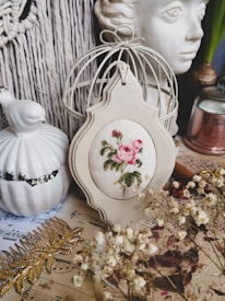A vintage-style still life arrangement featuring a cross-stitched floral design in a decorative frame, positioned among various items including a ceramic bird, a sculpted face, a white-wire birdcage, dried flowers, sheet music, and a golden leaf ornament. The setting suggests a cozy, artistic atmosphere.