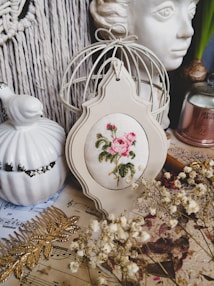 A vintage-style still life arrangement featuring a cross-stitched floral design in a decorative frame, positioned among various items including a ceramic bird, a sculpted face, a white-wire birdcage, dried flowers, sheet music, and a golden leaf ornament. The setting suggests a cozy, artistic atmosphere.