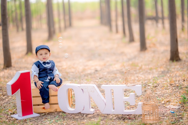 A young child dressed in a navy blue suit with a bow tie and cap is sitting on a wooden crate in a forested area, surrounded by trees. Large letters on the ground spell out the word 'ONE' with a red number one in front. Bubbles are floating around, and two decorative birdcages are gently placed nearby.