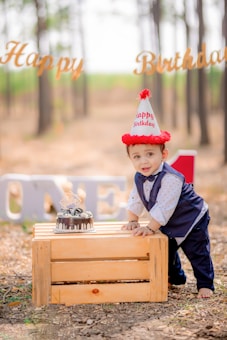 A young child in formal attire is leaning on a wooden crate outdoors. The child is wearing a party hat and standing next to a small birthday cake. In the background, there are trees and a 'Happy Birthday' sign, with large letters spelling out 'ONE'.