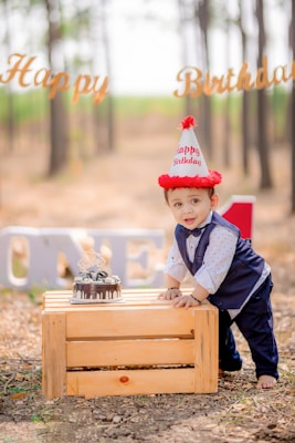 A young child in formal attire is leaning on a wooden crate outdoors. The child is wearing a party hat and standing next to a small birthday cake. In the background, there are trees and a 'Happy Birthday' sign, with large letters spelling out 'ONE'.