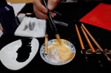 A close-up of an artist mixing gold pigment in a small dish with a brush. Nearby, there is another dish containing black ink and several brushes placed on a holder. A napkin with ink stains and a bottle of ink are also visible on the table. The focus on the mixing process suggests a preparation for a painting or calligraphy session.