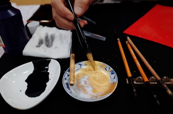 A close-up of an artist mixing gold pigment in a small dish with a brush. Nearby, there is another dish containing black ink and several brushes placed on a holder. A napkin with ink stains and a bottle of ink are also visible on the table. The focus on the mixing process suggests a preparation for a painting or calligraphy session.