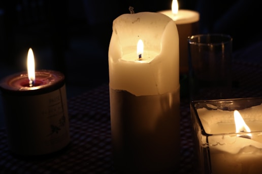 Several lit candles of different sizes and shapes are placed in a dimly lit environment. The central candle is tall and white, partially melted, casting a warm glow. A smaller, red wax candle in a glass jar is visible to the left, and another candle in a square glass holder is in the foreground. The soft, flickering flames create a cozy ambiance.