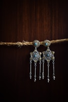 Two ornate earrings with intricate designs hang from a natural fiber rope. The earrings feature star-like patterns and are adorned with small turquoise stones and dangling chains. The background is a dark wood finish that contrasts with the metallic elements of the jewelry.