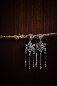 Two ornate earrings with intricate designs hang from a natural fiber rope. The earrings feature star-like patterns and are adorned with small turquoise stones and dangling chains. The background is a dark wood finish that contrasts with the metallic elements of the jewelry.