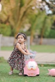 A young child in a floral dress is playing with a small pink ride-on toy with cartoon features, set outdoors on a grassy area with a background of blurred trees and a pathway.