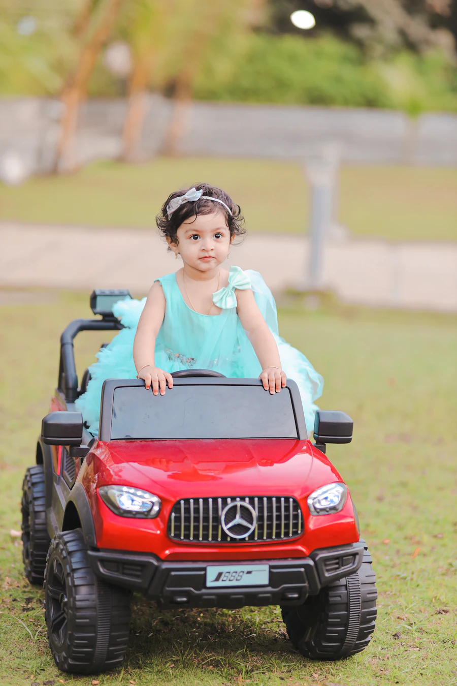 A little girl in a party dress driving a red ride-on Mercedes G-Wagon