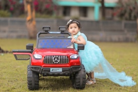 A young child wearing a fluffy, light blue dress is standing next to a red toy car designed to resemble a luxury vehicle. The child appears to be curious or playful, with one hand resting on the car. The background includes a grassy area and a blurred view of a building and trees.