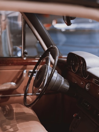 Interior of a vintage car featuring a classic steering wheel and dashboard. The seat and door panel have a beige leather finish with wood accents.