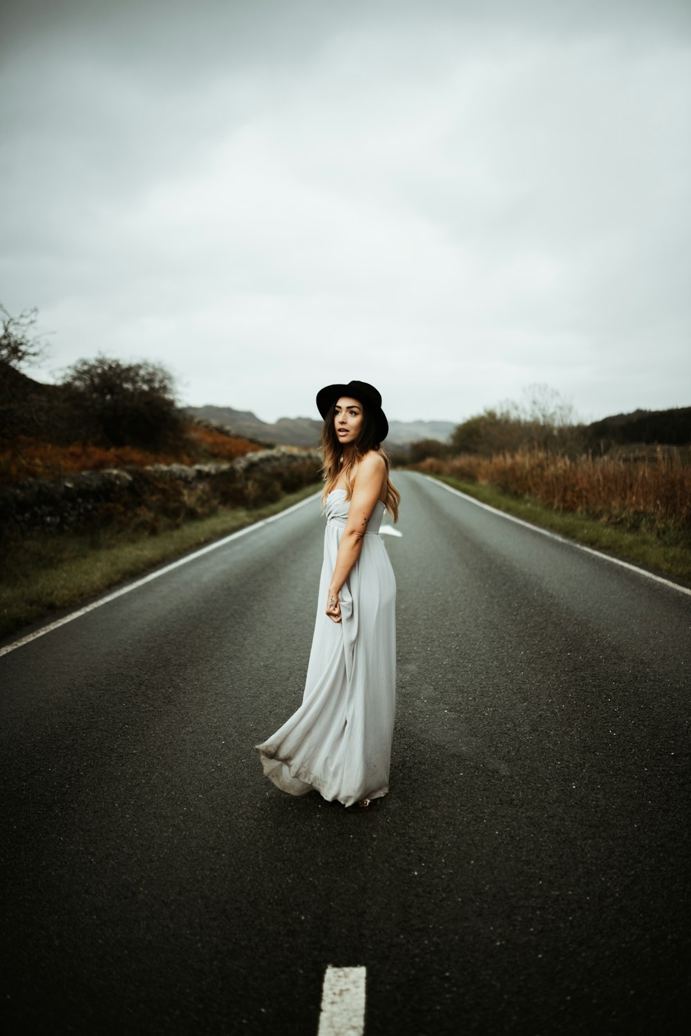a woman in a dress and hat standing on the side of a road