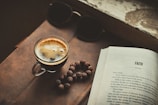 A close-up view of a glass cup filled with a dark, freshly brewed espresso placed on a leather surface. To the right is an open book with the title 'FAITH' on the page. There are a pair of black sunglasses and a beaded string next to the coffee cup.
