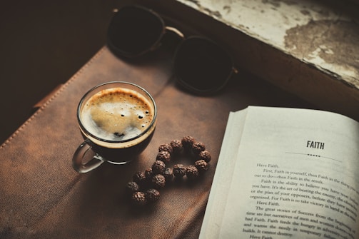 A close-up view of a glass cup filled with a dark, freshly brewed espresso placed on a leather surface. To the right is an open book with the title 'FAITH' on the page. There are a pair of black sunglasses and a beaded string next to the coffee cup.