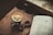 A close-up view of a glass cup filled with a dark, freshly brewed espresso placed on a leather surface. To the right is an open book with the title 'FAITH' on the page. There are a pair of black sunglasses and a beaded string next to the coffee cup.