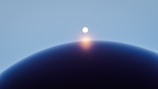 A celestial scene featuring a bright, glowing sphere hovering above the dark curve of a planet or moon. The soft gradient from deep blue to light blue in the background gives a sense of space and depth.