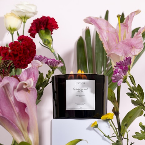 A black candle with a label titled 'Esencia' stands on a white pedestal surrounded by a variety of colorful flowers including red carnations, pink lilies, and yellow chrysanthemums. The candle emits a warm glow, contributing to the serene ambience.