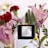 A black candle with a label titled 'Esencia' stands on a white pedestal surrounded by a variety of colorful flowers including red carnations, pink lilies, and yellow chrysanthemums. The candle emits a warm glow, contributing to the serene ambience.