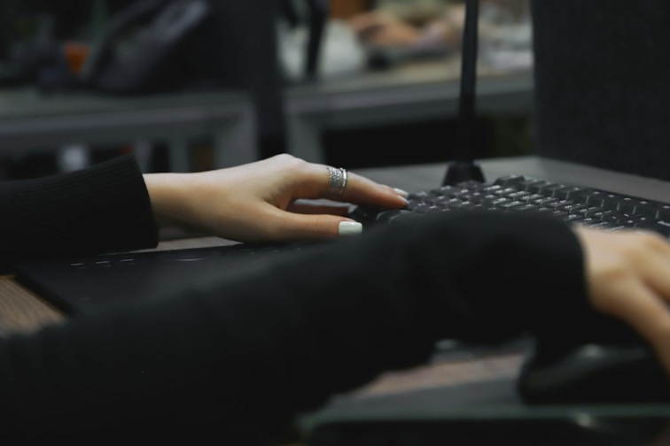 A close-up of a person's hands typing on a keyboard. One hand is partially visible with silver rings and painted nails, while the other hand is manipulating a computer mouse. The background is blurred, suggesting a typical office workspace or computer lab.