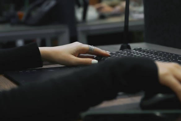 A close-up of a person's hands typing on a keyboard. One hand is partially visible with silver rings and painted nails, while the other hand is manipulating a computer mouse. The background is blurred, suggesting a typical office workspace or computer lab.