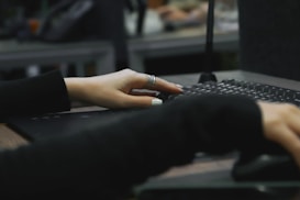 A close-up of a person's hands typing on a keyboard. One hand is partially visible with silver rings and painted nails, while the other hand is manipulating a computer mouse. The background is blurred, suggesting a typical office workspace or computer lab.