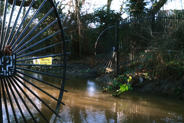 A black metal fence with circular designs is positioned next to a flowing stream in a wooded area. A warning sign reading 'Anti climb paint' is attached to the gate. The surrounding vegetation appears lush, with trees and scattered bushes.