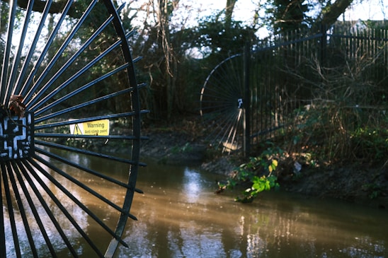 A black metal fence with circular designs is positioned next to a flowing stream in a wooded area. A warning sign reading 'Anti climb paint' is attached to the gate. The surrounding vegetation appears lush, with trees and scattered bushes.