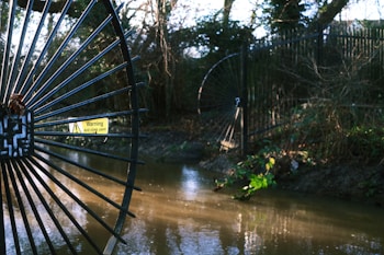 A black metal fence with circular designs is positioned next to a flowing stream in a wooded area. A warning sign reading 'Anti climb paint' is attached to the gate. The surrounding vegetation appears lush, with trees and scattered bushes.