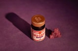 A small jar with a copper-colored lid labeled 'Dad Grass' is placed against a deep purple background. Next to the jar, there is a neatly stacked pile of dark red gummy candies shaped like cubes, casting a shadow.