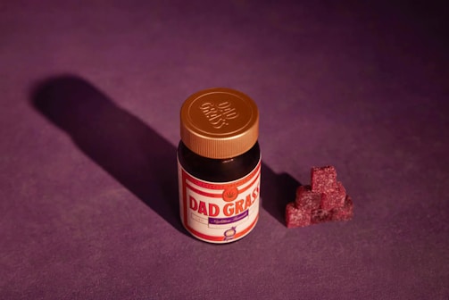 A small jar with a copper-colored lid labeled 'Dad Grass' is placed against a deep purple background. Next to the jar, there is a neatly stacked pile of dark red gummy candies shaped like cubes, casting a shadow.