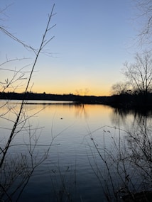 A calm lake reflecting the soft hues of a sunset. Bare branches frame the scene in the foreground, while sparse trees line the opposite shore. The sky transitions from a soft blue to warm oranges near the horizon.