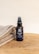 A bottle of beard oil with a dark label and pump dispenser is placed on a wooden surface next to folded towels. The label indicates it is enriched with vitamin E, argan oil, and macadamia oil.