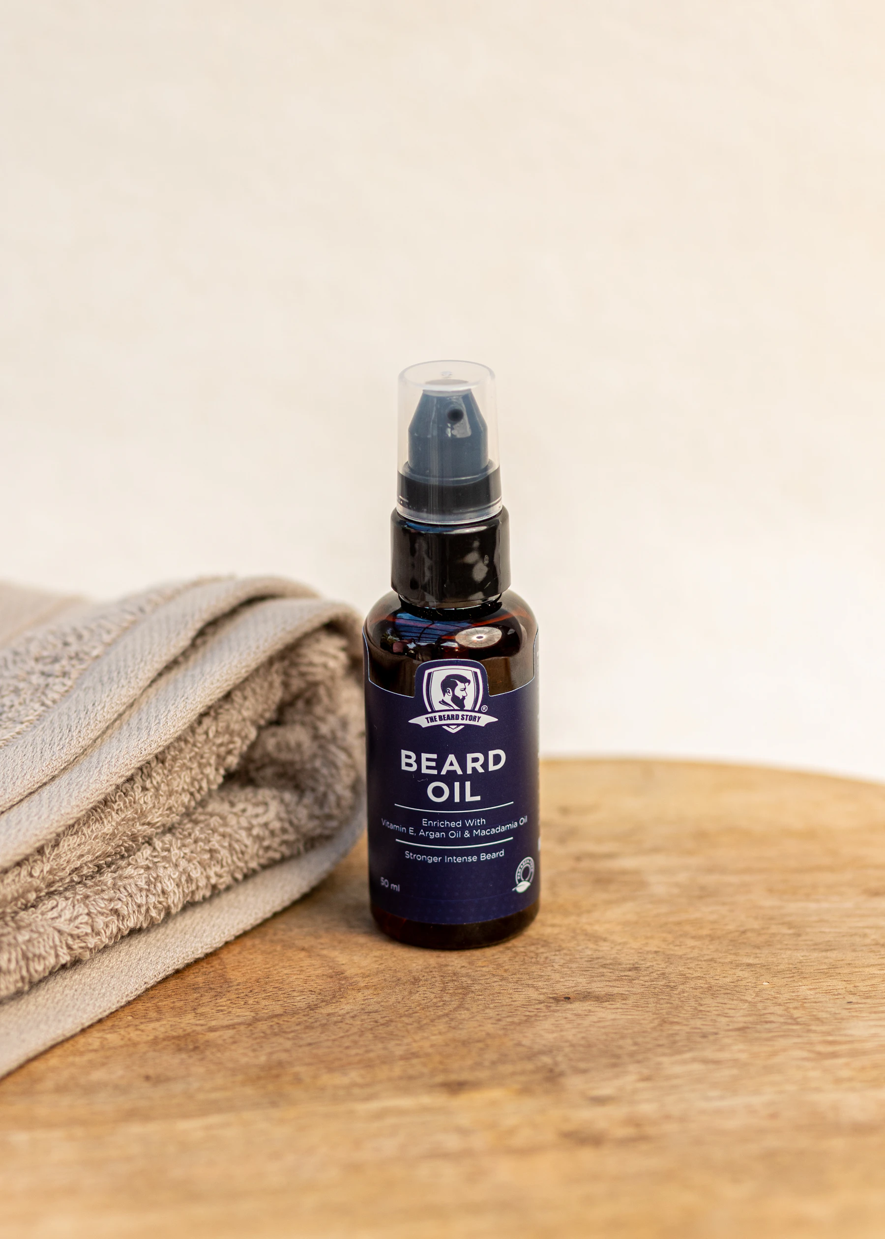 An amber pump bottle of beard oil standing on a round wooden board beside a folded gray towel