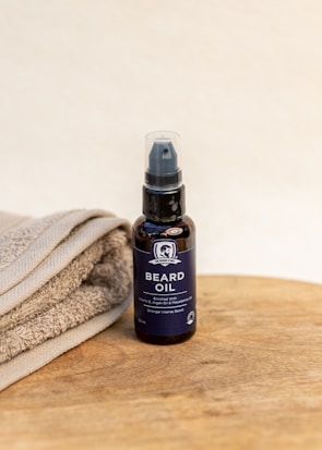 A bottle of beard oil with a dark label and pump dispenser is placed on a wooden surface next to folded towels. The label indicates it is enriched with vitamin E, argan oil, and macadamia oil.
