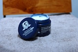 A blue container of The Beard Story Ultimate Men Daily Hair Cream is placed on a textured beige surface. The lid is off and leaning against the container, revealing creamy white contents inside. The background includes a wooden element, and the label on the container specifies its purpose and characteristics, such as being paraben-free.
