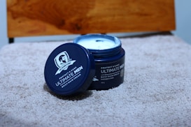 A blue container of The Beard Story Ultimate Men Daily Hair Cream is placed on a textured beige surface. The lid is off and leaning against the container, revealing creamy white contents inside. The background includes a wooden element, and the label on the container specifies its purpose and characteristics, such as being paraben-free.