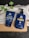Two blue bottles for hair care products, one labeled as a conditioner and the other as a shampoo, are placed on a folded white towel. There is also a green leafy plant visible beside the products. The items are set against a dark, textured surface, and the bottles prominently display branding for keratin and biotin products designed for men.
