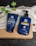 Two blue bottles for hair care products, one labeled as a conditioner and the other as a shampoo, are placed on a folded white towel. There is also a green leafy plant visible beside the products. The items are set against a dark, textured surface, and the bottles prominently display branding for keratin and biotin products designed for men.