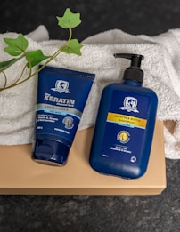 Two blue bottles for hair care products, one labeled as a conditioner and the other as a shampoo, are placed on a folded white towel. There is also a green leafy plant visible beside the products. The items are set against a dark, textured surface, and the bottles prominently display branding for keratin and biotin products designed for men.