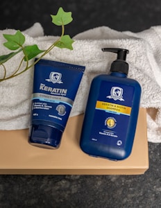 Two blue bottles for hair care products, one labeled as a conditioner and the other as a shampoo, are placed on a folded white towel. There is also a green leafy plant visible beside the products. The items are set against a dark, textured surface, and the bottles prominently display branding for keratin and biotin products designed for men.