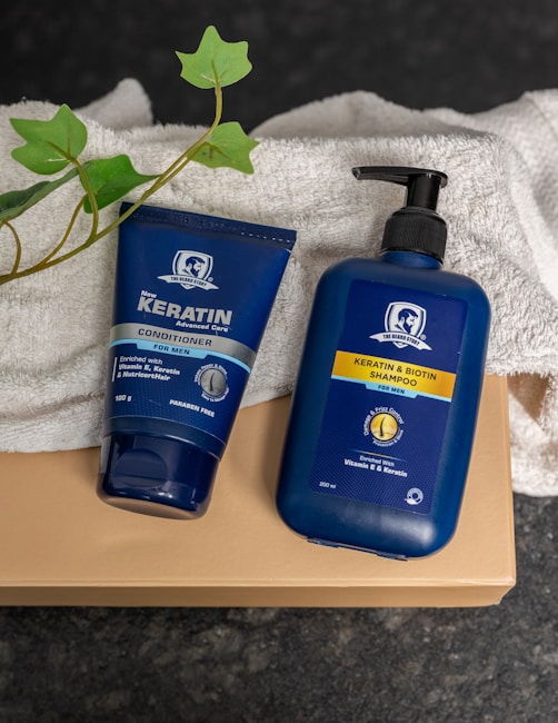 Two blue bottles for hair care products, one labeled as a conditioner and the other as a shampoo, are placed on a folded white towel. There is also a green leafy plant visible beside the products. The items are set against a dark, textured surface, and the bottles prominently display branding for keratin and biotin products designed for men.