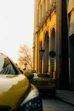 Yellow taxis are parked on a city street, with one prominently visible in the foreground. The warm glow of the setting or rising sun casts a golden hue on a tall building with arched windows. A few street signs and a lone tree without leaves add to the urban scene.
