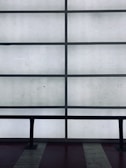 A grid of frosted glass panels is framed by dark metal bars, creating a minimalist architectural pattern. In front of the panels, a dark railing runs horizontally across the lower part of the image.