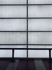 A grid of frosted glass panels is framed by dark metal bars, creating a minimalist architectural pattern. In front of the panels, a dark railing runs horizontally across the lower part of the image.