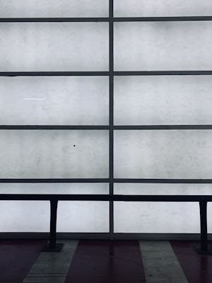A grid of frosted glass panels is framed by dark metal bars, creating a minimalist architectural pattern. In front of the panels, a dark railing runs horizontally across the lower part of the image.