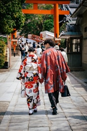 Two people dressed in traditional Japanese attire walk along a stone path lined with trees and buildings. In the background, a traditional torii gate stands prominently, leading to a busy area filled with more people.