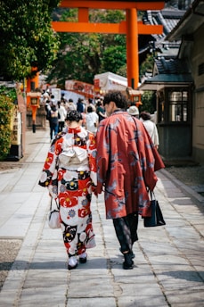 Two people dressed in traditional Japanese attire walk along a stone path lined with trees and buildings. In the background, a traditional torii gate stands prominently, leading to a busy area filled with more people.