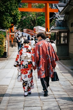 Two people dressed in traditional Japanese attire walk along a stone path lined with trees and buildings. In the background, a traditional torii gate stands prominently, leading to a busy area filled with more people.