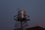 A metallic water tank is elevated on a black lattice stand against a dimly lit evening sky. The tank reflects a subtle amount of light, creating a contrast with the darker surroundings. The roof of a building is partially visible at the base with a weather vane or antenna on top.