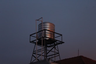A metallic water tank is elevated on a black lattice stand against a dimly lit evening sky. The tank reflects a subtle amount of light, creating a contrast with the darker surroundings. The roof of a building is partially visible at the base with a weather vane or antenna on top.