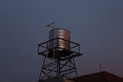 A metallic water tank is elevated on a black lattice stand against a dimly lit evening sky. The tank reflects a subtle amount of light, creating a contrast with the darker surroundings. The roof of a building is partially visible at the base with a weather vane or antenna on top.